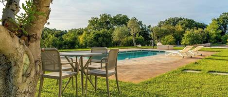 A pool area with a table and chairs, surrounded by grass and trees.