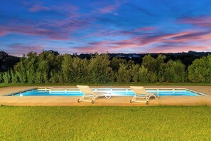 Una piscina rectangular con dos sillas de descanso mirando al agua, rodeada de césped y una hilera de árboles al fondo.