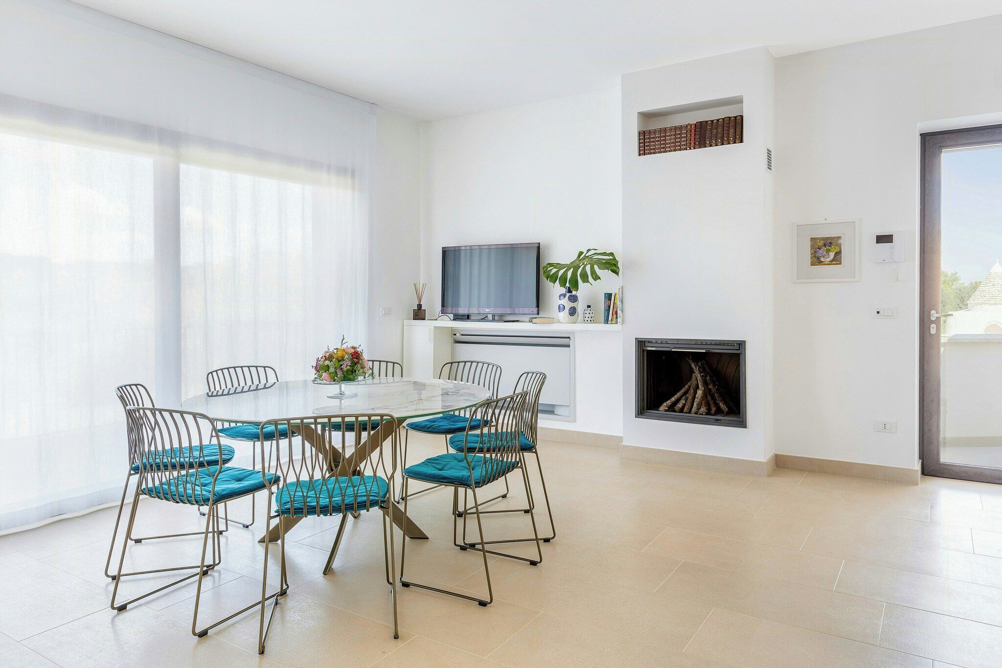 A modern dining area with a round table and six chairs, a flat-screen TV, a fireplace, and a large window with sheer curtains.