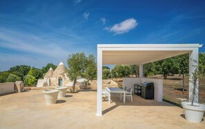 Un espacio al aire libre techado con una parrilla y asientos, rodeado de construcciones de piedra y árboles.