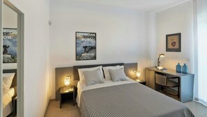 A hotel room with a bed, bedside tables, a desk, and a chair. There are posters on the walls and a framed certificate on the desk.
