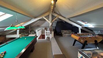A room with a pool table, foosball table, and a billiards table. There is a ceiling fan, a chandelier, and a window providing natural light.