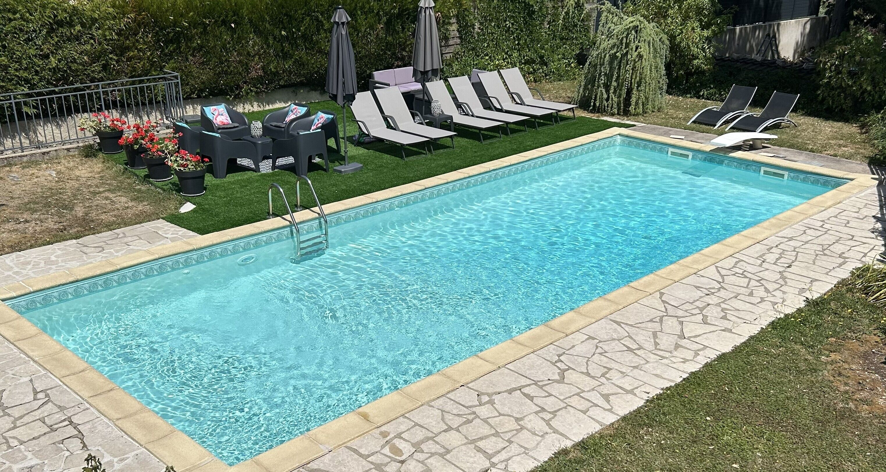 A rectangular swimming pool with a stone border, surrounded by a grassy area and a row of lounge chairs.