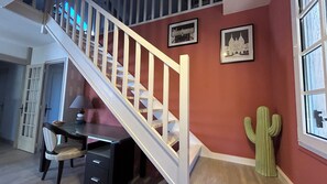 A staircase with white balusters leading to a landing, a desk with a chair, a cactus decoration, and two framed pictures on the wall.