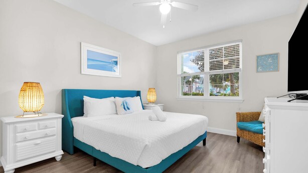A bedroom with a bed, white nightstands, a ceiling fan, a window with blinds, and a view of the outside. - Runaway Beach Club Disney Retreat (Kissimmee)