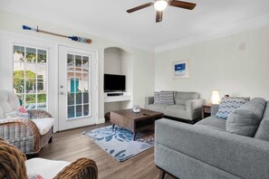 Ein Wohnzimmer mit grauem Sofa, einem Holztisch, einem Fernseher auf einem Standfuß und einem Deckenventilator.