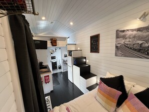 A compact hotel room with a kitchenette, a small bathroom, a bed with colorful pillows, and a wall-mounted painting.