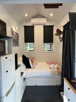 A compact bedroom with a bed, a small desk, a heater, and a window with a view of greenery.