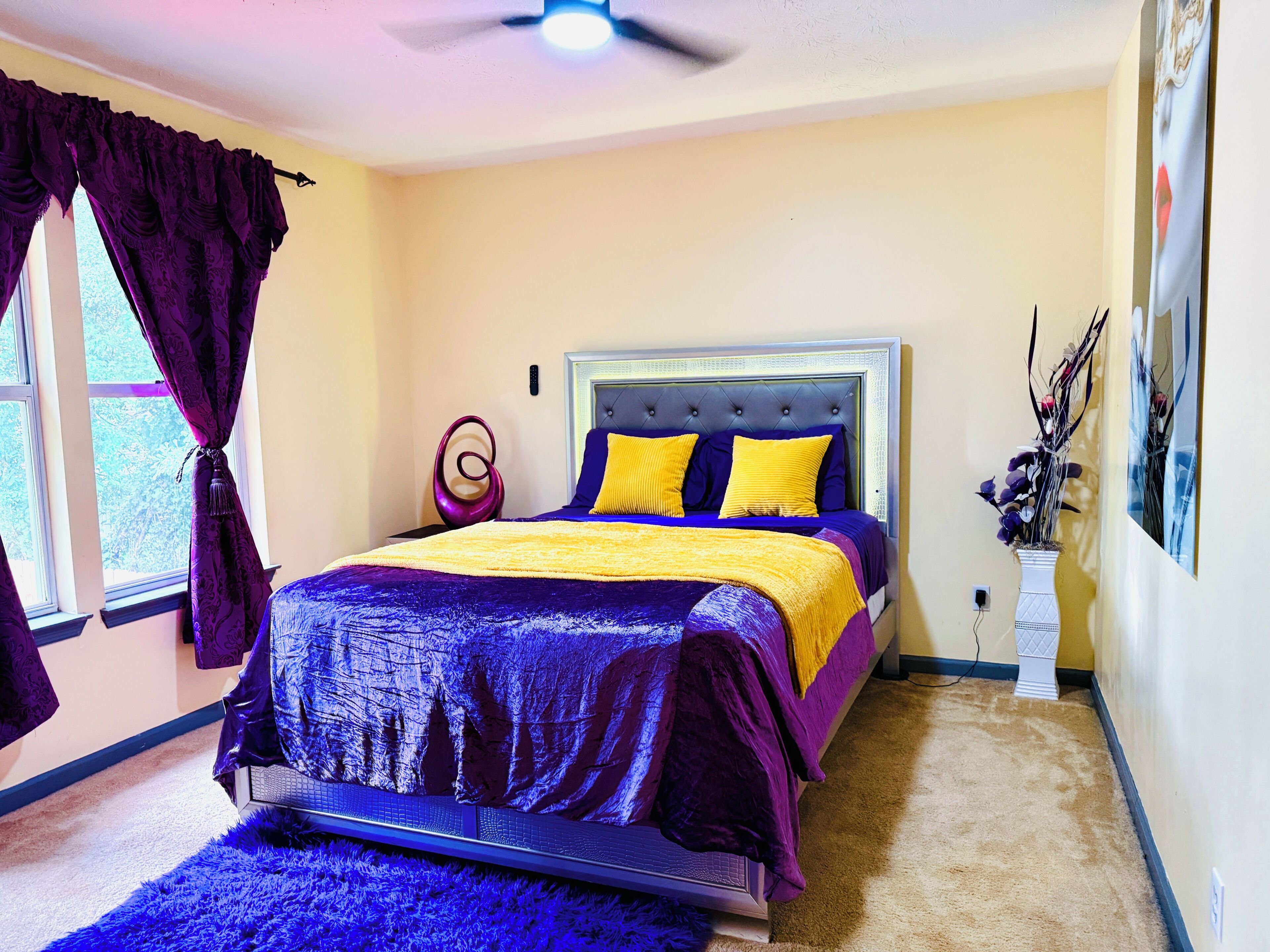 A bedroom with a bed, curtains, a vase with flowers, and a ceiling fan.