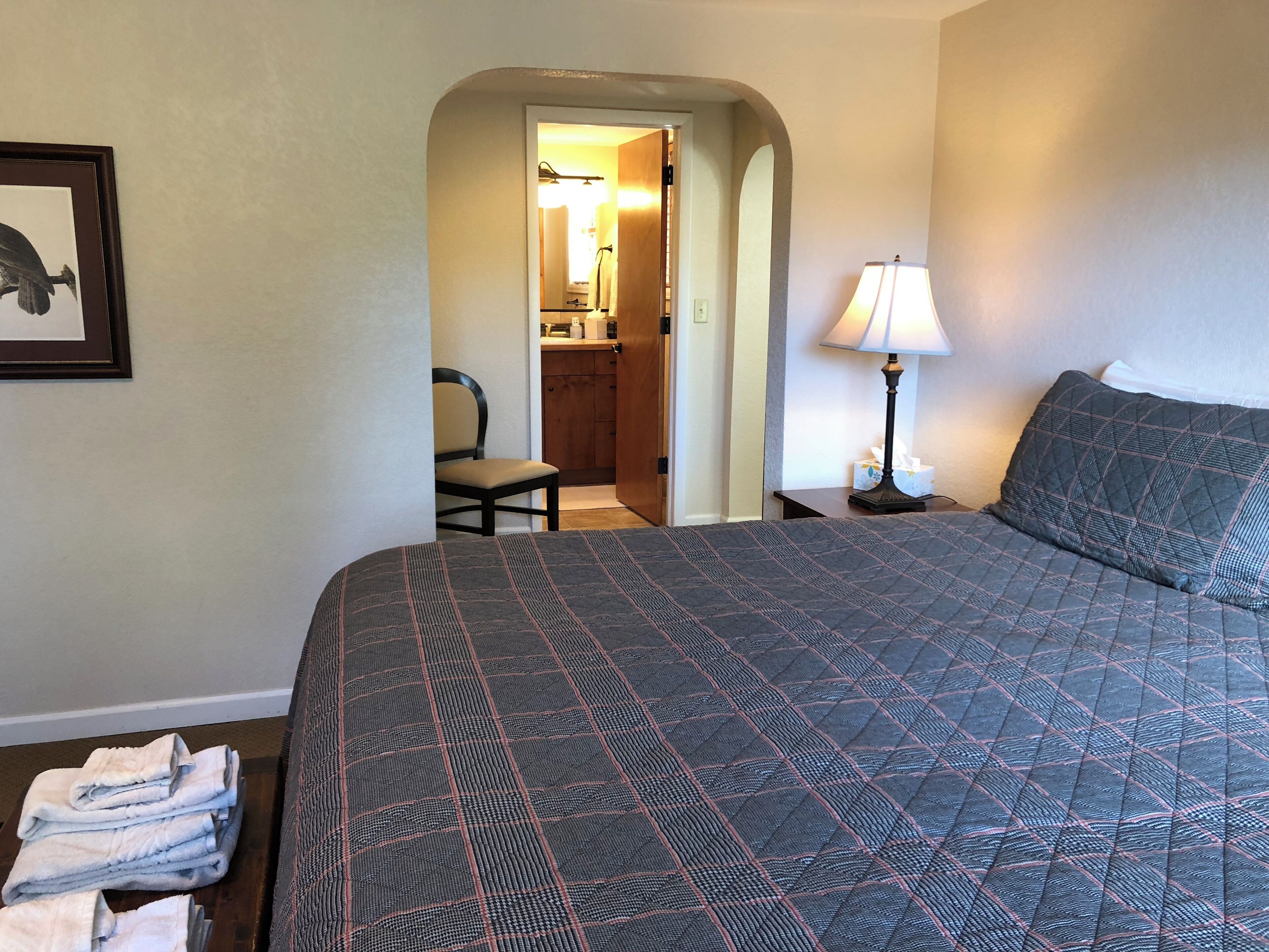 A hotel room with a large bed, a chair, a nightstand with a lamp, a framed picture on the wall, and a doorway leading to a bathroom.