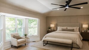 A bedroom with a large bed, a ceiling fan, a window with a view of trees, a chair, and a small table with a lamp. - Luxury Estate Weddings • Retreats • Group Stays (Akron)
