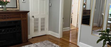 A room with hardwood flooring, a fireplace, a large mirror, and a door leading to another room.