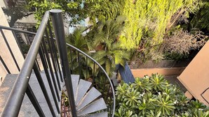 Una escalera de caracol con barandillas metálicas que conduce a un balcón con plantas en macetas y vistas a un árbol.