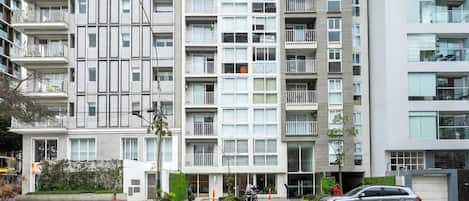 A multi-story residential building with balconies, a gated entrance, and a parking area with vehicles.
