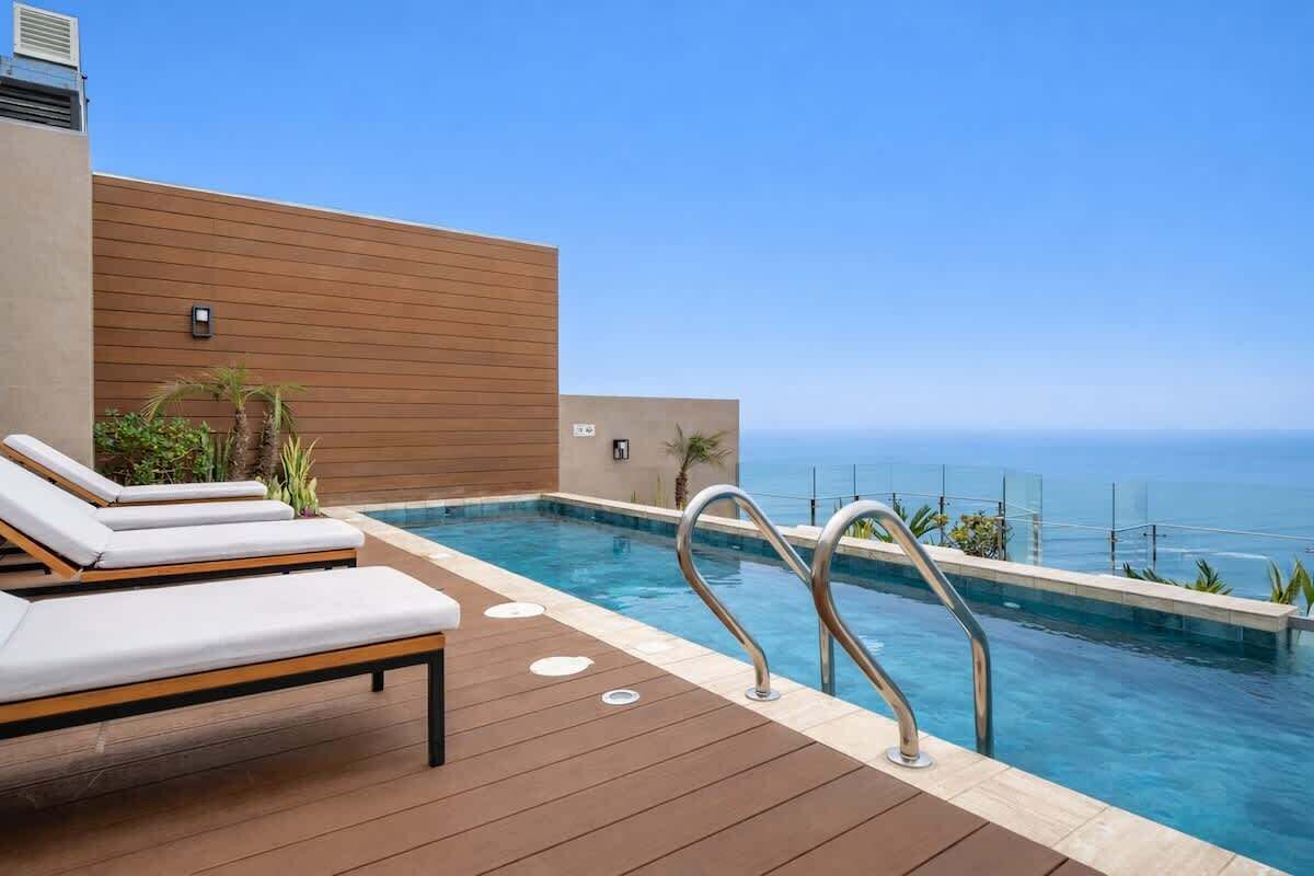 A rooftop infinity pool with lounge chairs, a wooden deck, and a clear view of the ocean.
