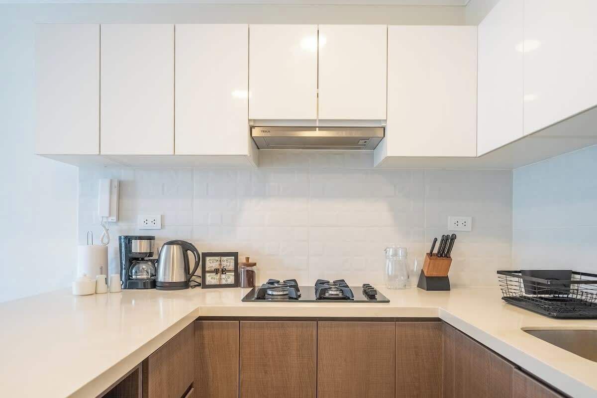A modern kitchen with white cabinets, a stainless steel range hood, and a countertop with a coffee maker, kettle, and utensils.