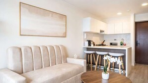 A modern living room with a beige sofa, a round wooden coffee table, and a kitchen area with white cabinets and bar stools. - Sight 1601 · Luxe King Bed 1BR •Rooftop Pool, Gym, AC & Parking (Lima)