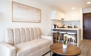 A modern living room with a beige sofa, a round wooden coffee table, and a kitchen area with white cabinets and bar stools.