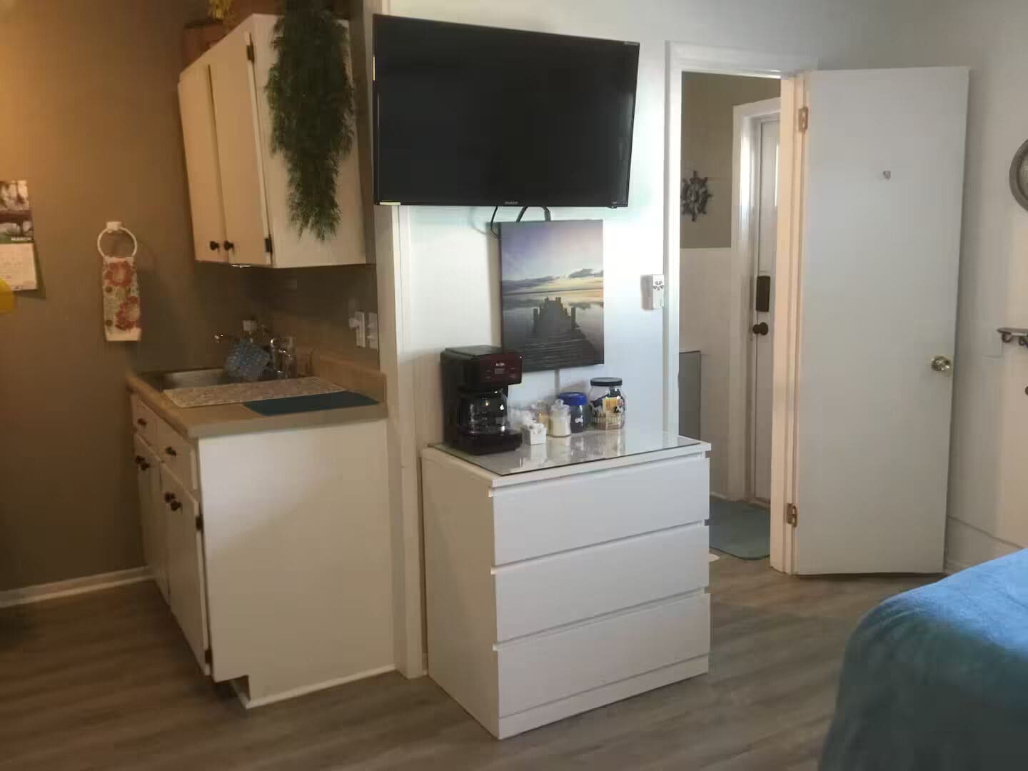 A compact room with a kitchenette, a flat-screen TV mounted above the counter, and a door leading to another room.