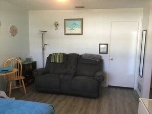 Ein Zimmer mit einem dunkelgrauen Sofa, einem kleinen Holzsessel, einem Schreibtisch mit Stuhl, einer Stehlampe und einer Tür.