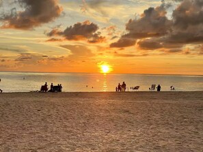 Eine Strandansicht mit untergehender Sonne über dem Wasser, Silhouetten von Personen und Liegestühlen.
