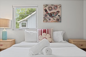 A neatly made bed with a rolled towel, a window with a view of a house, and a wall with a floral artwork.