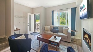 Ein modernes Wohnzimmer mit Sofa, Couchtisch und Sesseln. Ein Kamin und ein an der Wand befestigter Fernseher sind ebenfalls vorhanden.