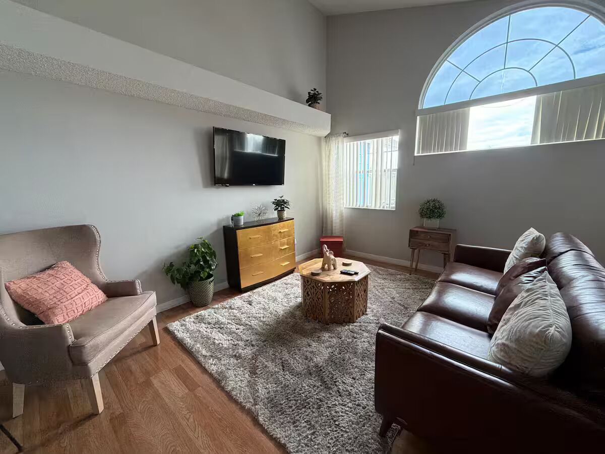 Ein Wohnzimmer mit Wand-Fernseher, braunem Ledersofa, Couchtisch aus Holz und einem flauschigen, grauen Teppich.