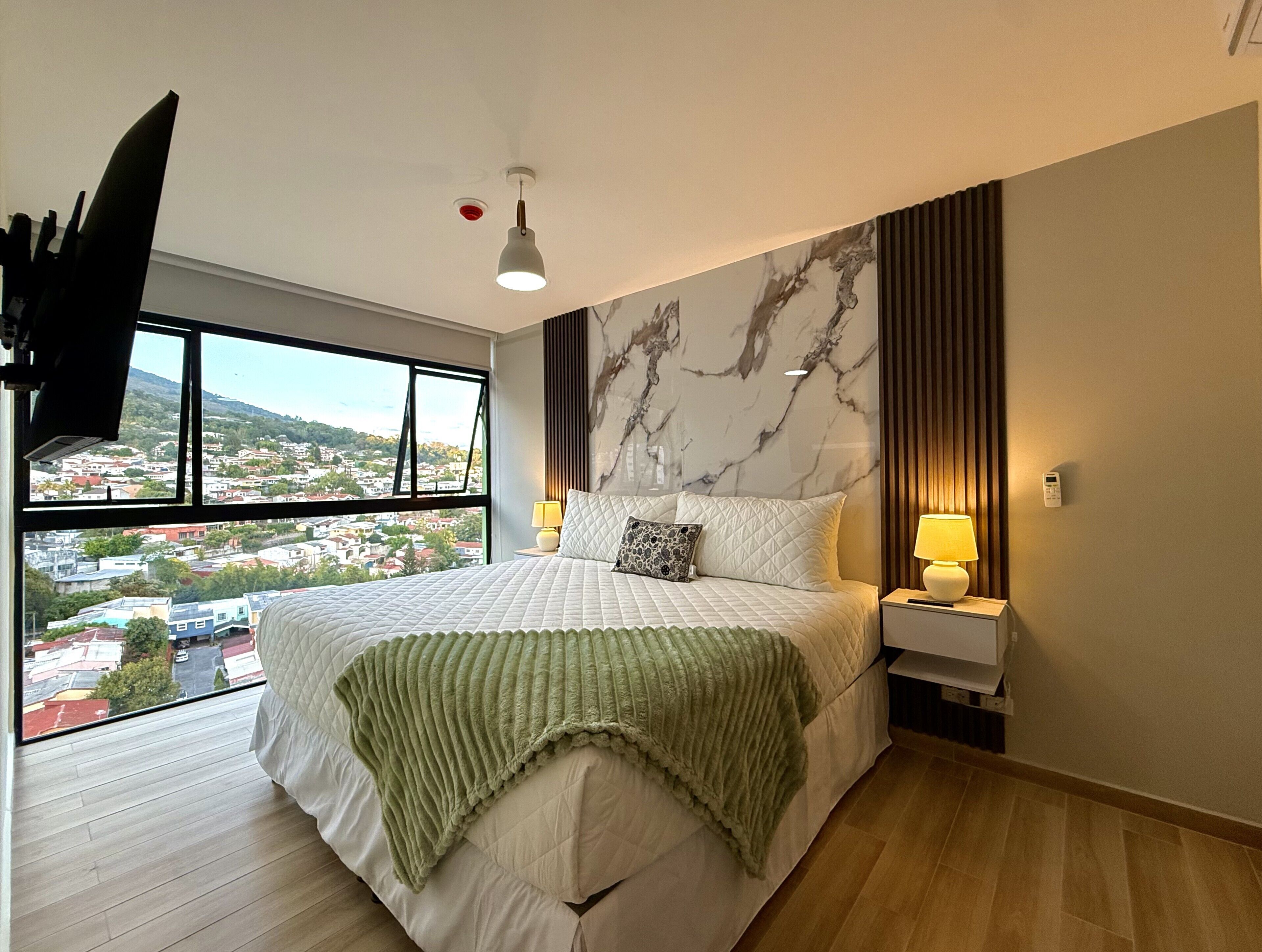 Habitación de hotel con una cama grande, vistas a la ciudad, un televisor de pantalla plana y una lámpara moderna.