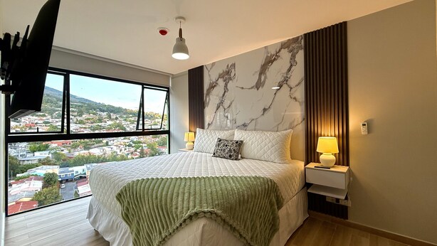 A hotel room with a large bed, a view of the city, a flat-screen TV, and a modern light fixture. - Cozy 1 bedroom apartment with AC in San Salvador (San Salvador)