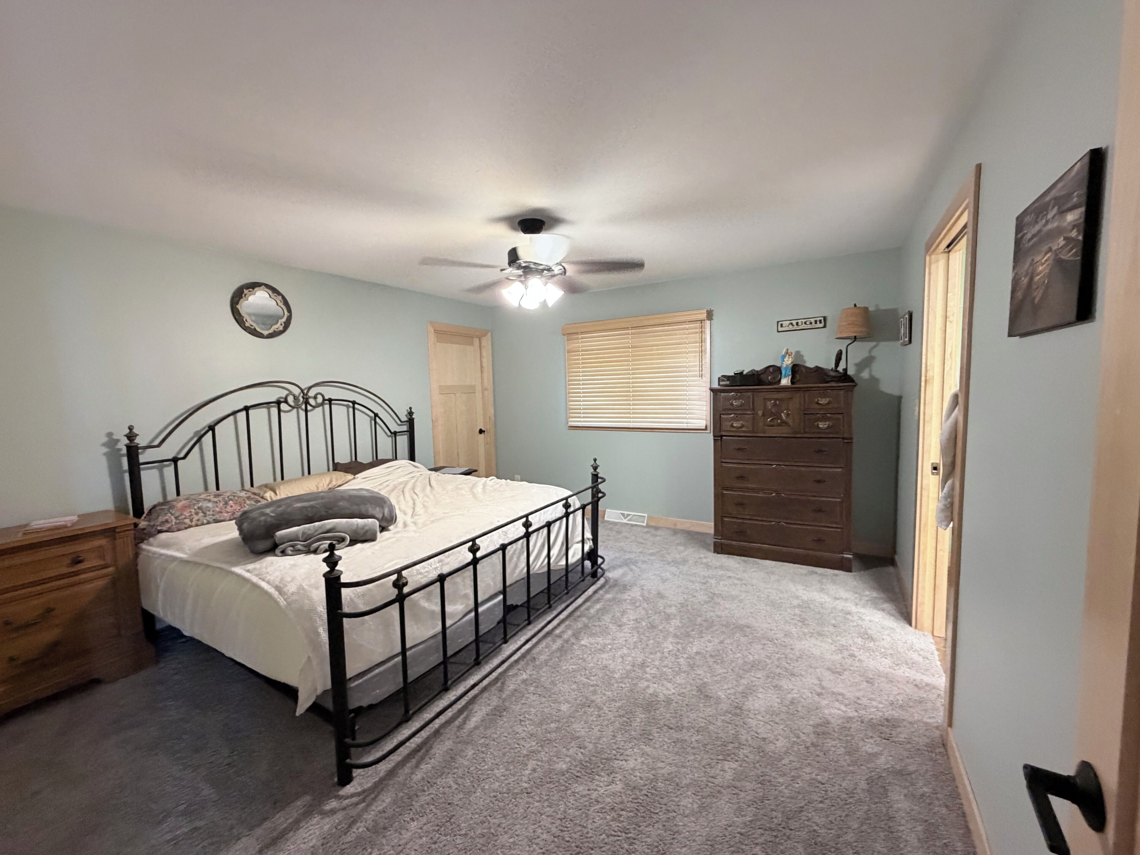 A bedroom with a bed, a dresser, a ceiling fan, a window with blinds, and a door.