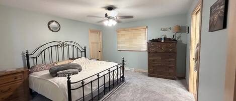 A bedroom with a bed, a dresser, a ceiling fan, a window with blinds, and a door.