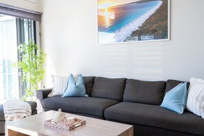 Ett vardagsrum med en grå soffa, ett soffbord med dekorativa föremål, ett stort fönster med persienner och en inramad tavla på väggen.