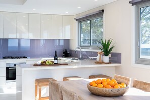A modern kitchen with white cabinetry, a central island, and a dining area with wicker chairs. - Allure: Panoramic beachfront views. Sunrise views over the ocean at Hyams Beach (Hyams Beach)