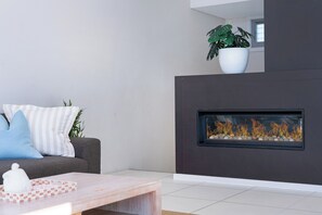 Ett modernt vardagsrum med en grå soffa, randiga kuddar, ett vitt bord med en rutig duk, en krukväxt och en öppen spis med en glaslucka.