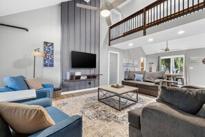 A spacious living room with a flat-screen TV, a dining area with a table and chairs, and a staircase leading to an upper level.