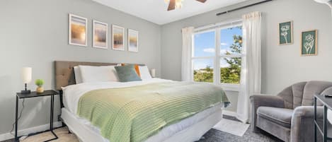 Un dormitorio con cama, escritorio, silla, ventilador de techo y ventana con cortinas.