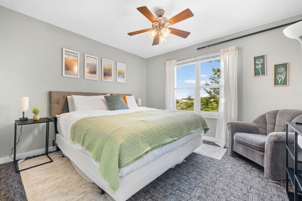 A bedroom with a bed, a desk, a chair, a ceiling fan, and a window with curtains.
