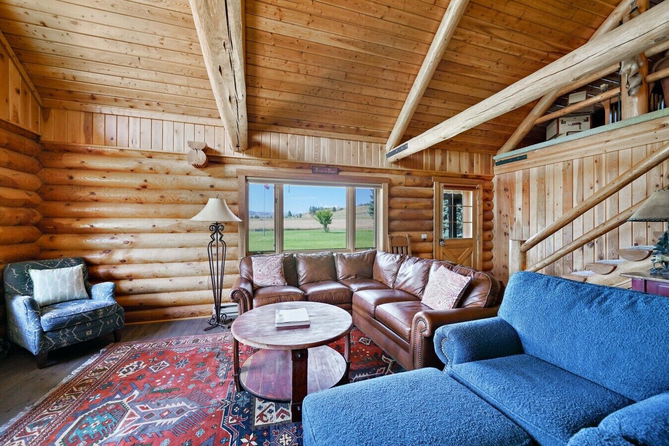 A log cabin interior with a leather sofa, a patterned rug, and a view of the outdoors.
