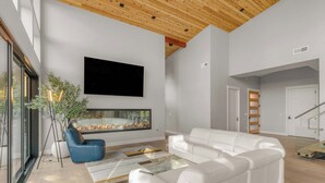 Uma sala de estar moderna com lareira, sofás brancos, uma poltrona azul e uma televisão grande.