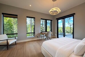 Ein Schlafzimmer mit einem großen Bett, einem Schreibtisch und einem Stuhl. Drei große Fenster bieten einen Blick ins Freie.