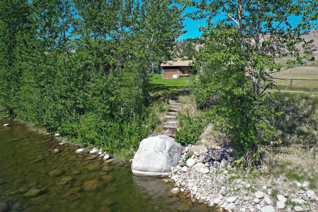A natural landscape with a clear stream, a wooden bridge, and a building in the background.