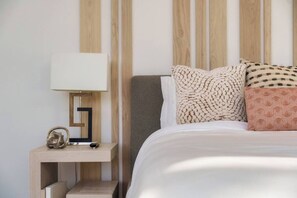 Ein ordentlich bezogenes Bett mit weißer Decke und gemusterten Kissen, ein Nachttisch mit Lampe und ein hölzernes Kopfteil.