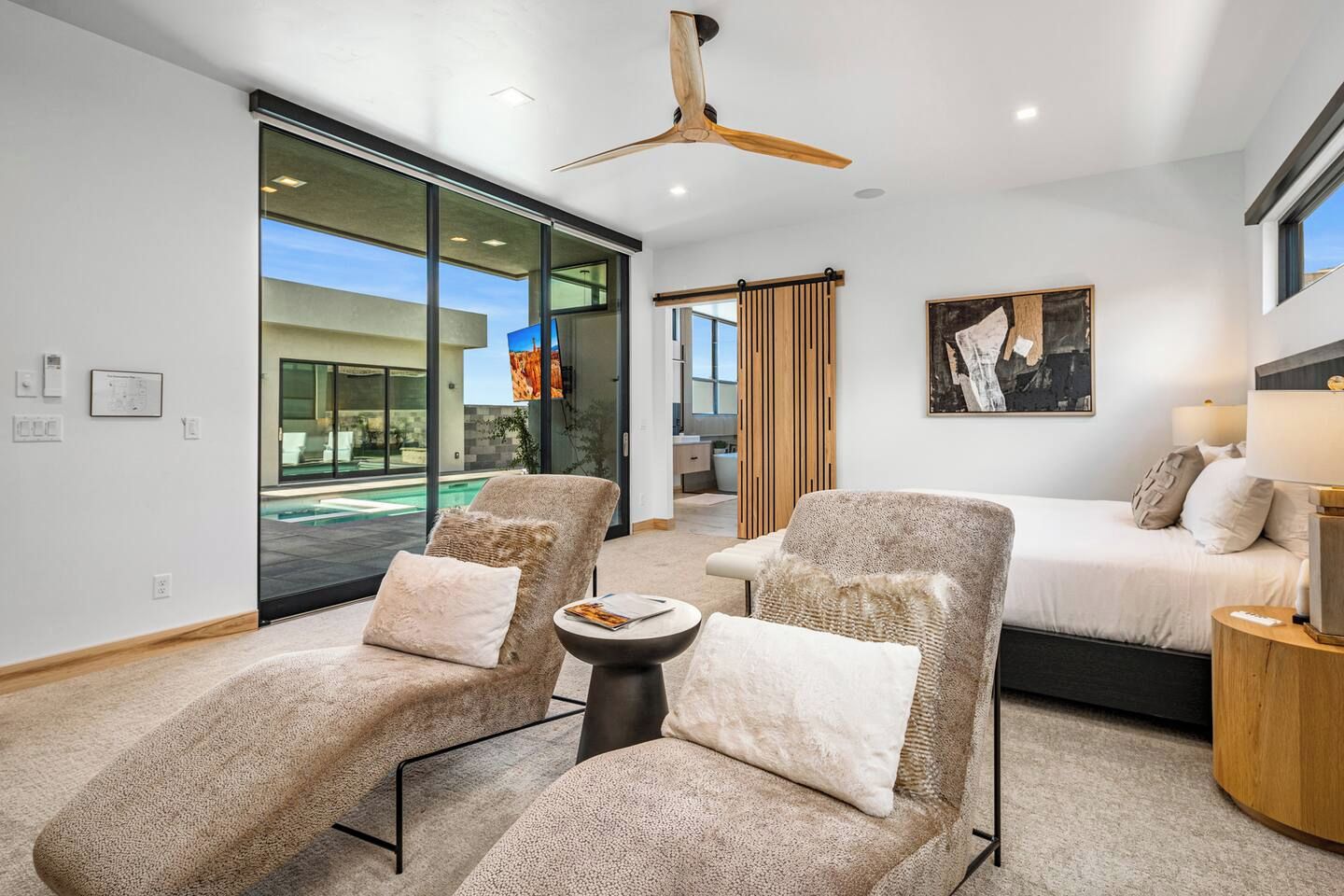 A modern hotel room with a large sliding glass door leading to a pool, a bed with a headboard, two armchairs, a round coffee table, and a ceiling fan.