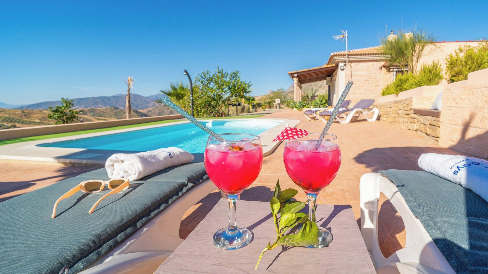 A poolside setting with two drinks, a towel, and lounge chairs.