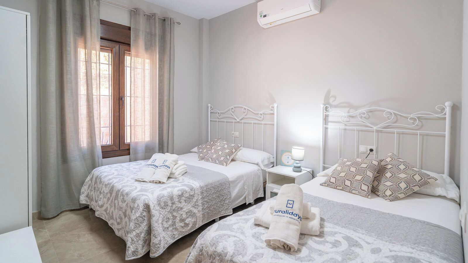 A hotel room with two beds, a nightstand, a lamp, and a window with curtains.