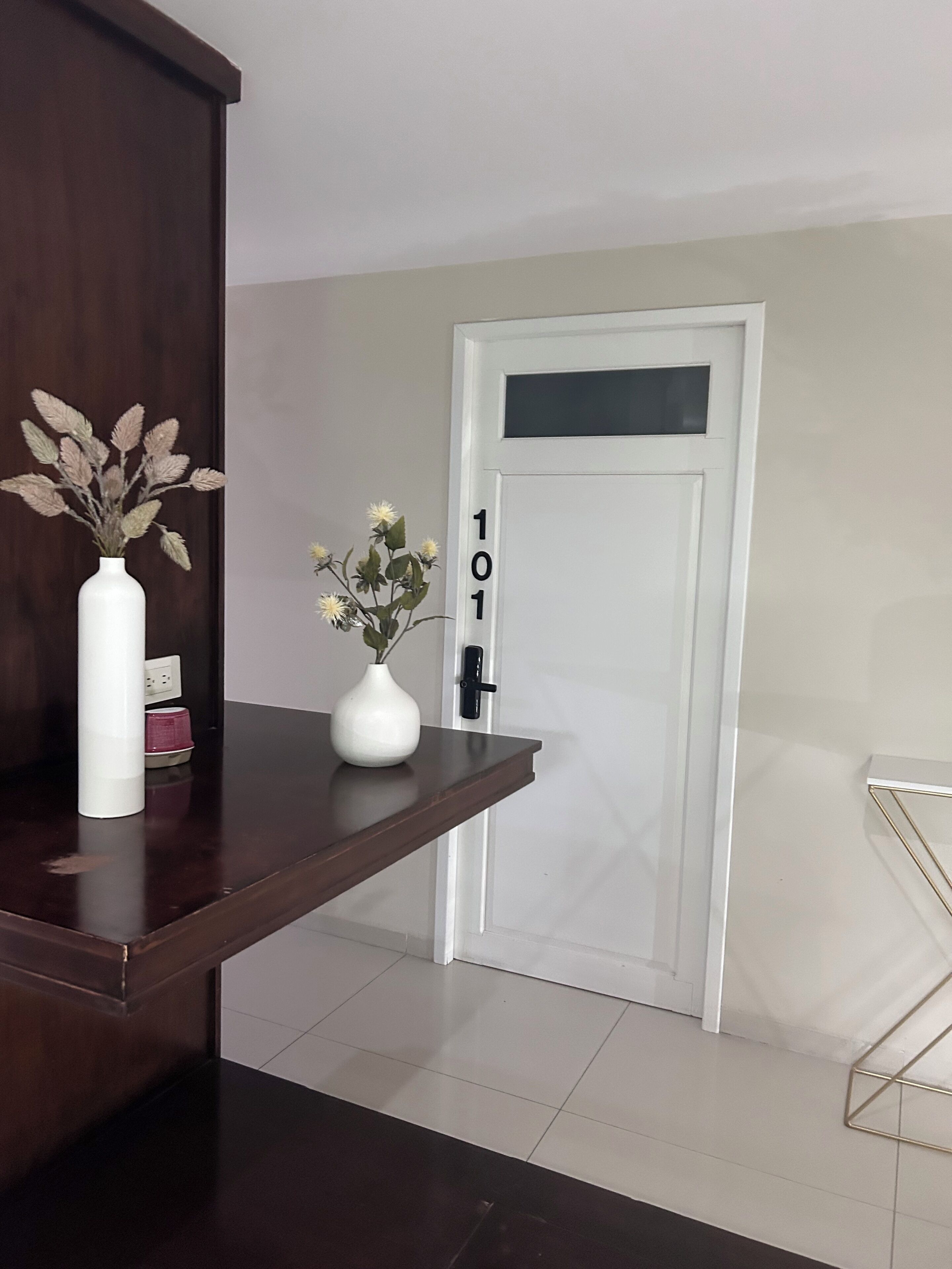 A room with a dark wooden shelf, a white vase with dried plants, a white vase with flowers, and a door with the number 101.