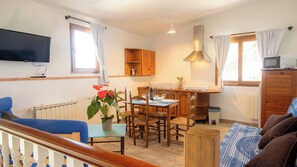 A room with a dining table, chairs, a television, a kitchen area, and a window with curtains.
