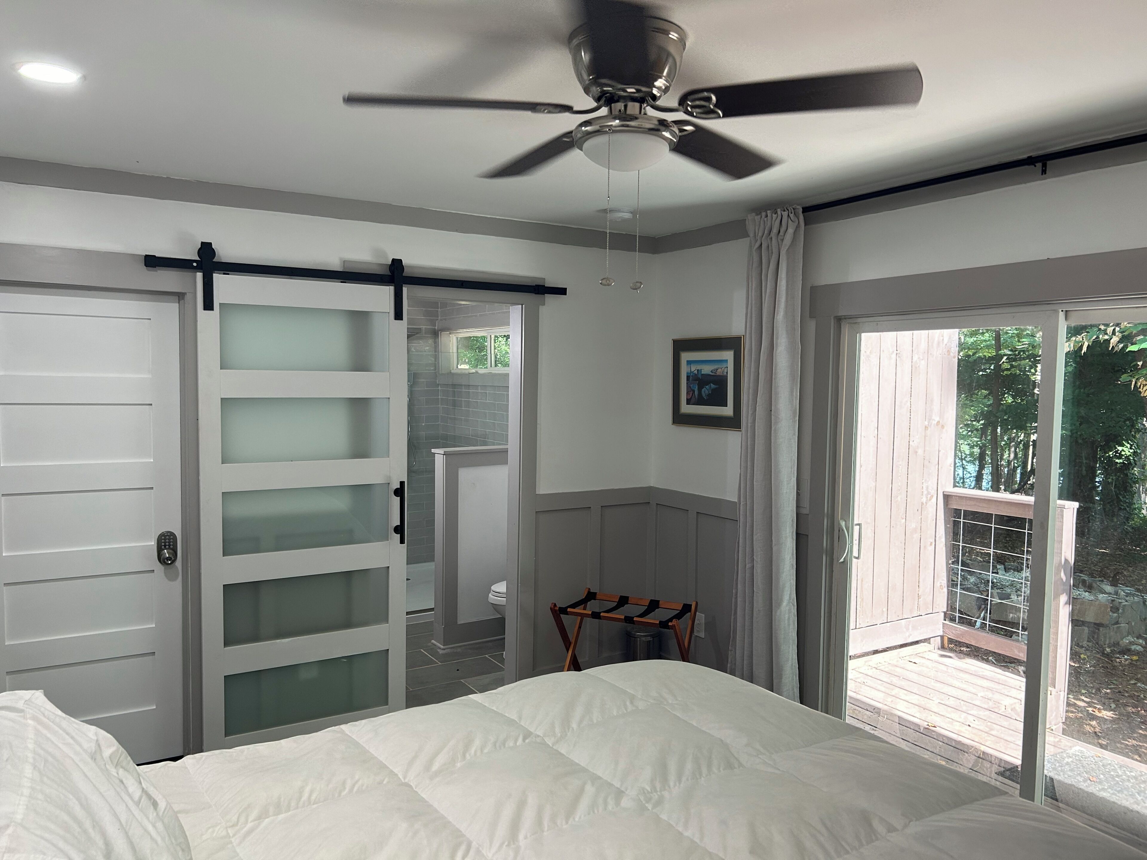 A bedroom with a bed, a ceiling fan, a sliding glass door leading to a balcony, and a bathroom with a toilet and sink.