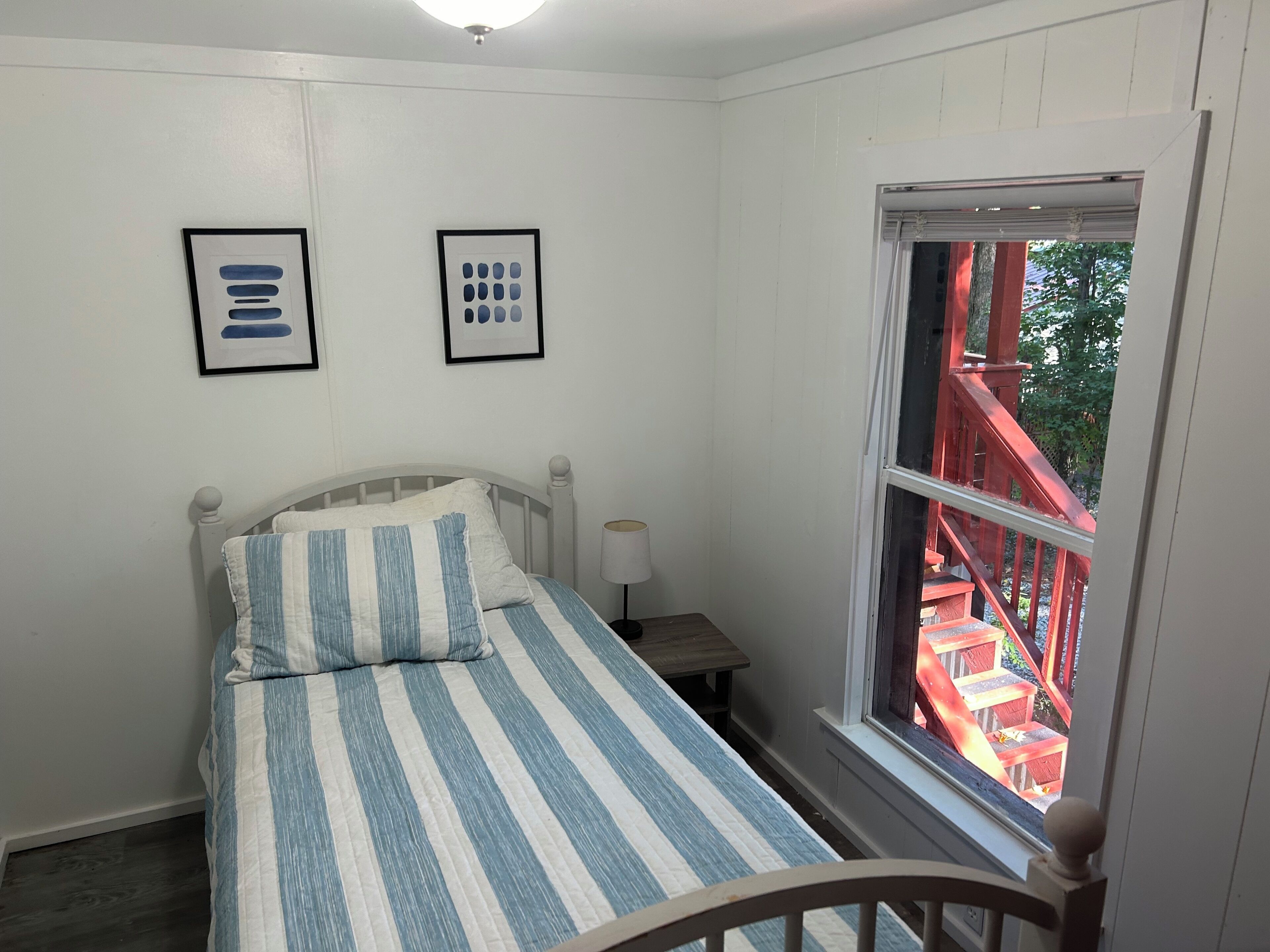 A bedroom with a bed, a nightstand, a window with a view of a red staircase, and two framed pictures on the wall.